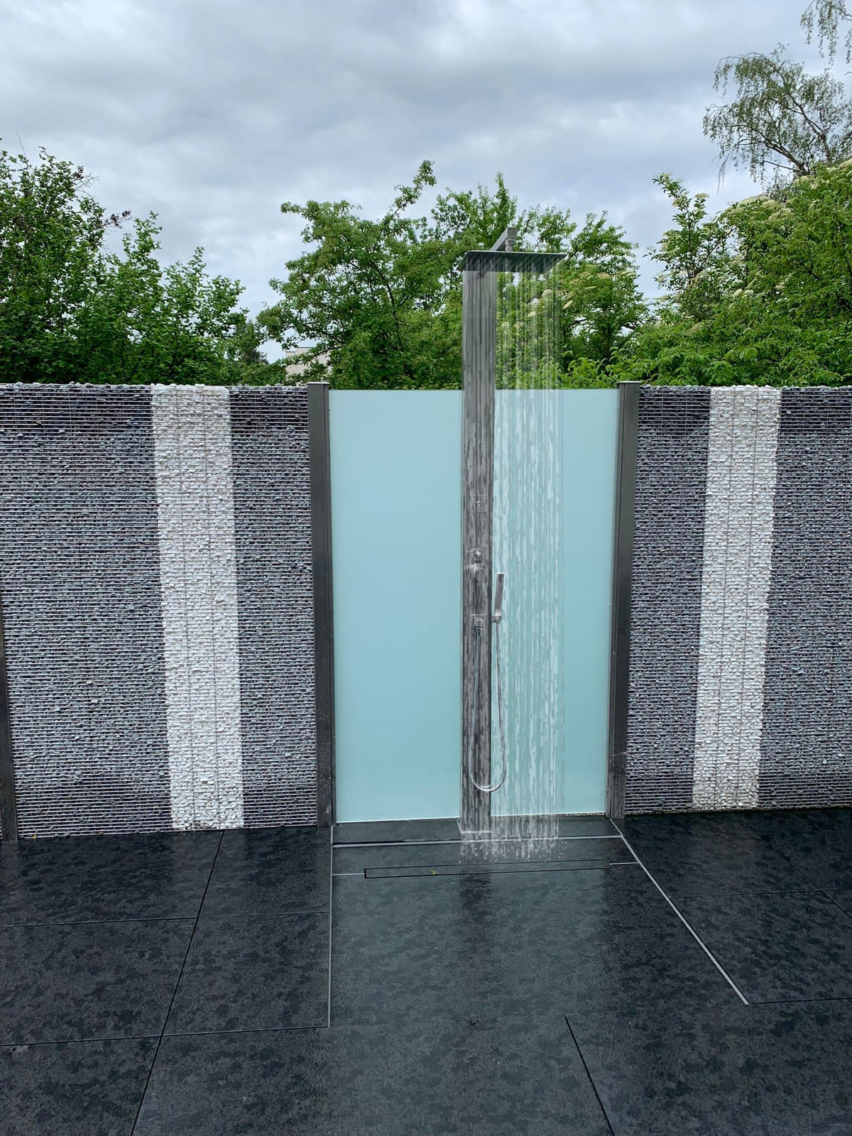 Edelstahl-Gartendusche in Betrieb – Wasser aus dem Regenwald-Duschkopf vor Milchglas und Steinkorb-Sichtschutz