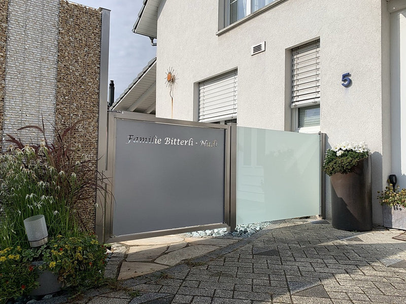 Sichtschutz-Element mit gelasertem Familiennamen als Schriftzug, daneben Steinkorb und Milchglas