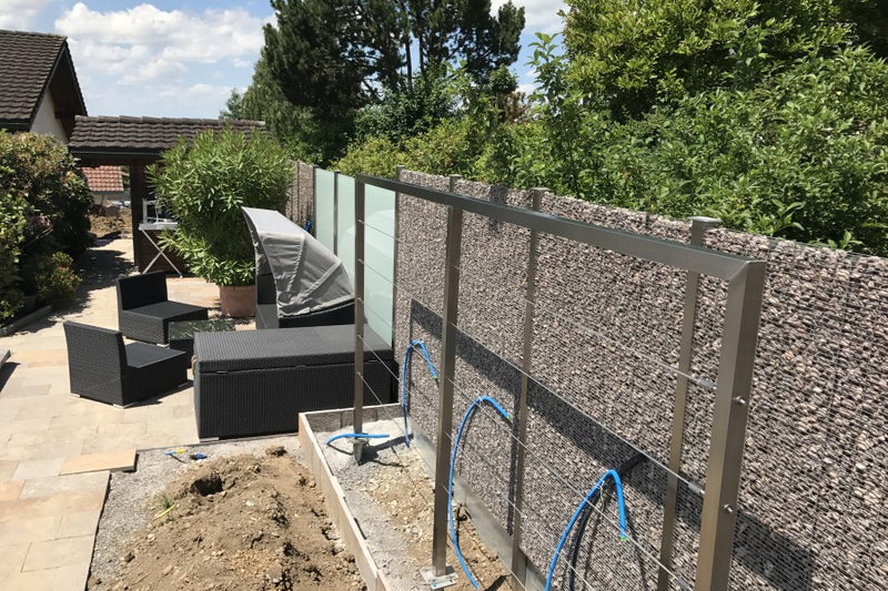 Sicht-, Wind- und Schallschutz aus Gabionen und VSG-Milchglas, mit Rankhilfe aus Edelstahlseilen – Bettwiesen